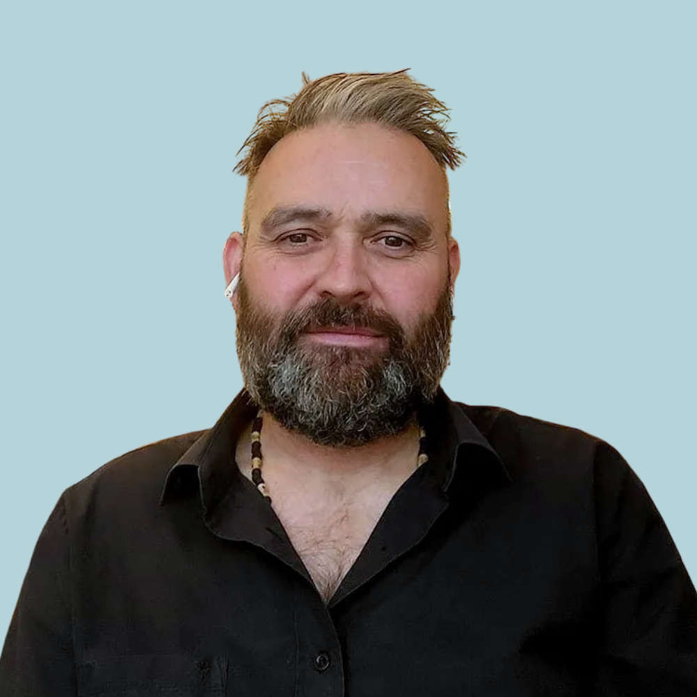 Man with beard and shaved head, wearing a black shirt and a necklace, against a light blue background.