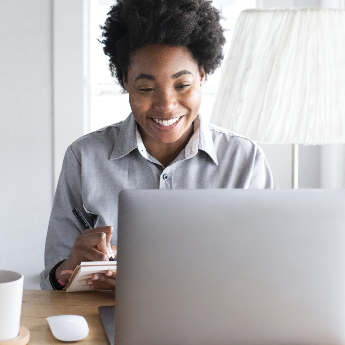 Woman smiling, taking notes while using a laptop at a desk with a lamp in the background.