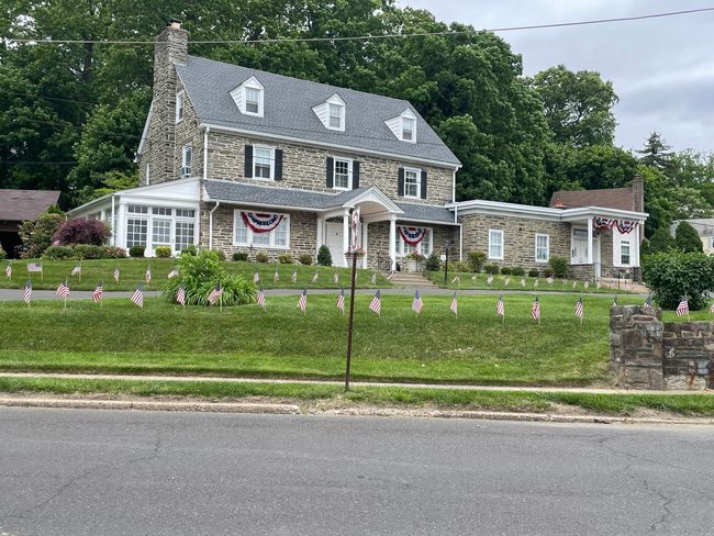 Stone house with gray roof, white porch, and patriotic bunting on a landscaped lawn by the road