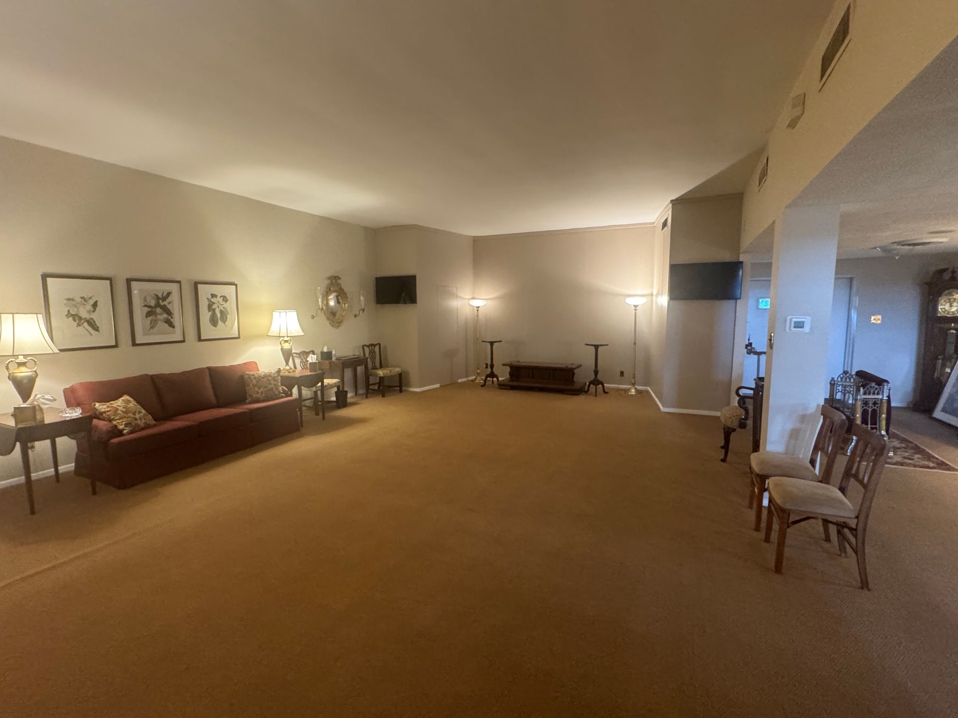 Spacious beige living room with a red sofa, framed wall art, lamps, and chairs along the right wall.