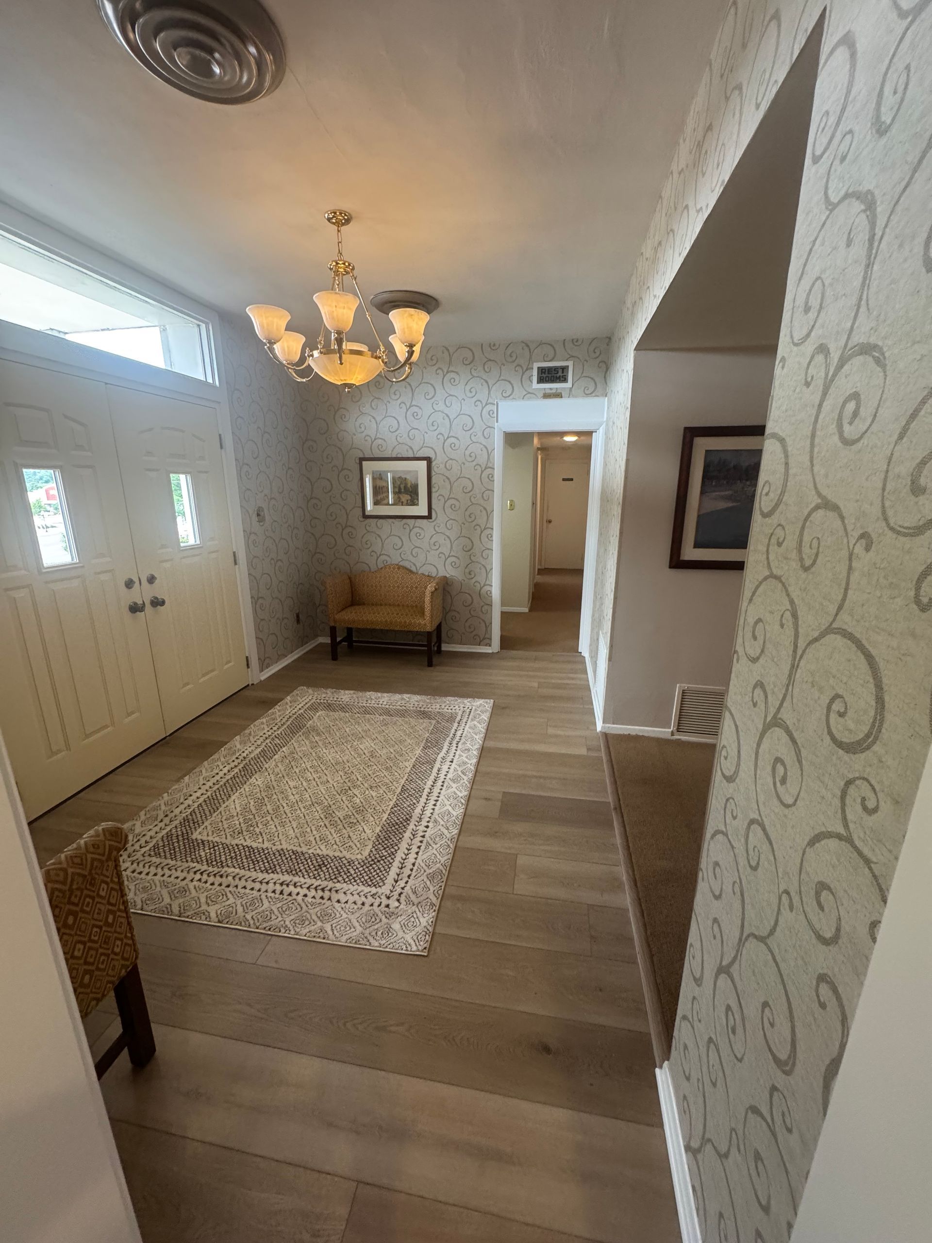 Bright foyer with white doors, patterned rug, chandelier, bench, and hallway leading to a doorway.