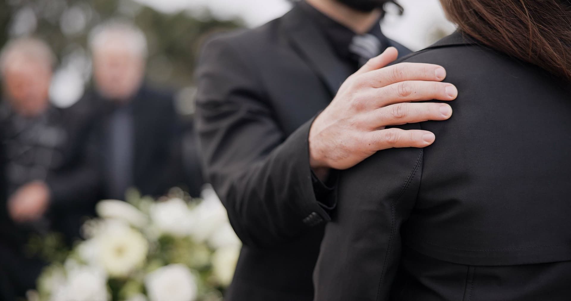 Mourners embrace at a funeral, with white flowers blurred in the background.