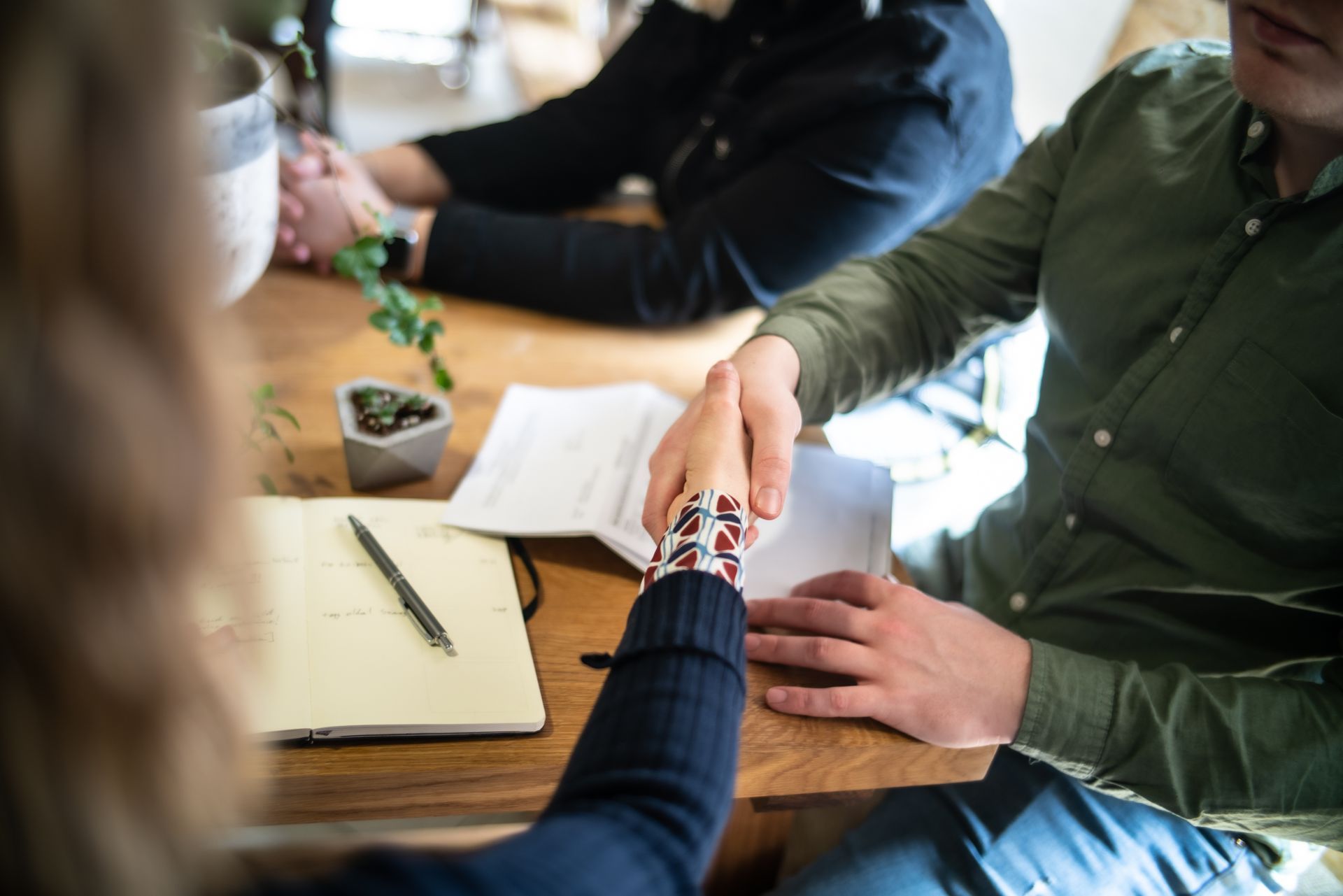 Two people shake hands over a wooden desk with a notebook and a small plant, while a third person sits nearby. Two people shake hands over a wooden desk with a notebook and a small plant, while a third person sits nearby.
