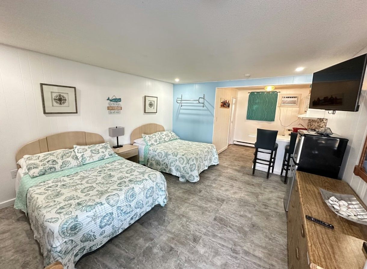 Two beds with patterned bedding in a hotel room with light walls, blue accents, a television, and a kitchenette.