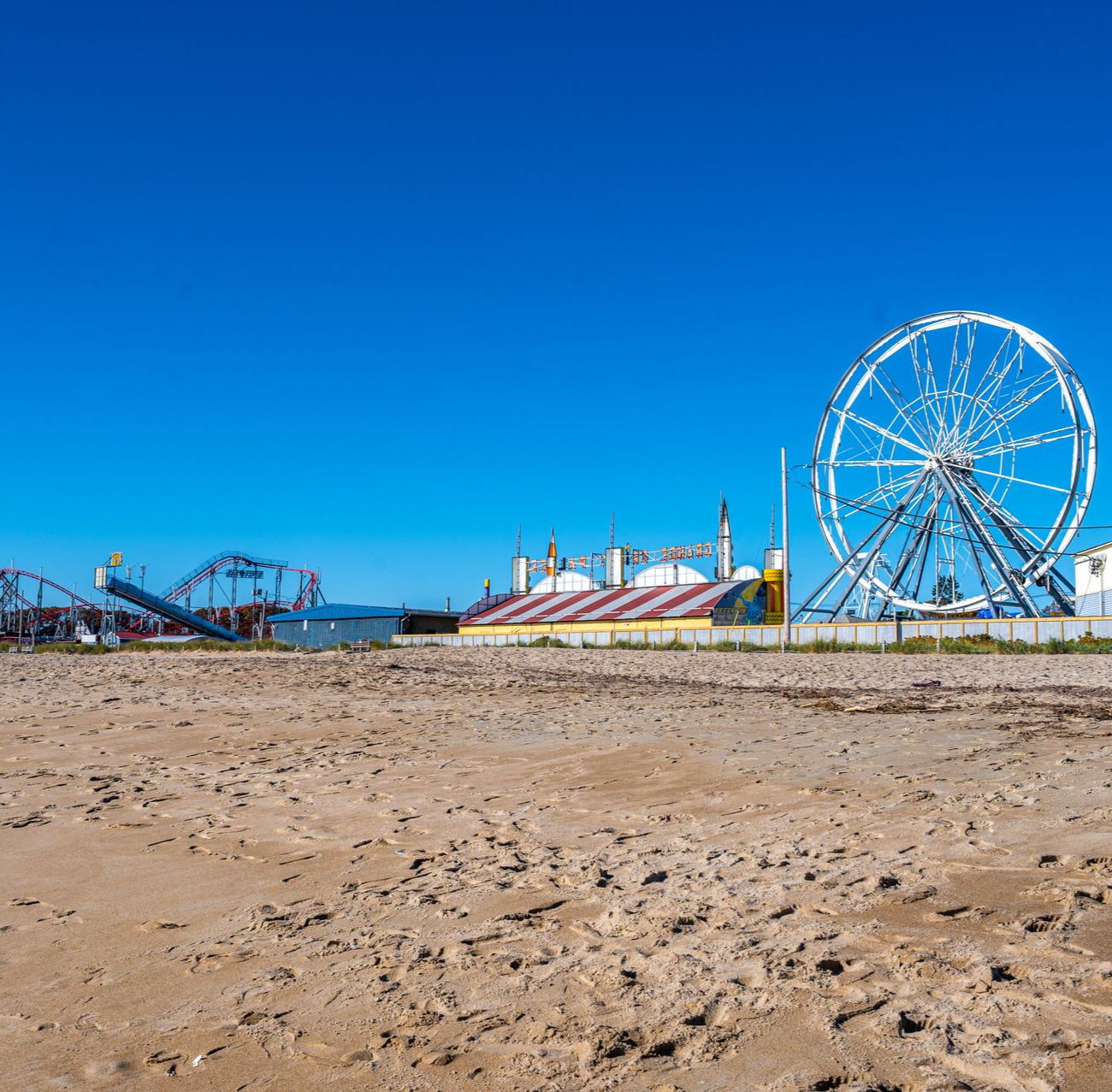 A sandy beach stretches toward a boardwalk with an amusement park featuring a large Ferris wheel under a clear blue sky.
