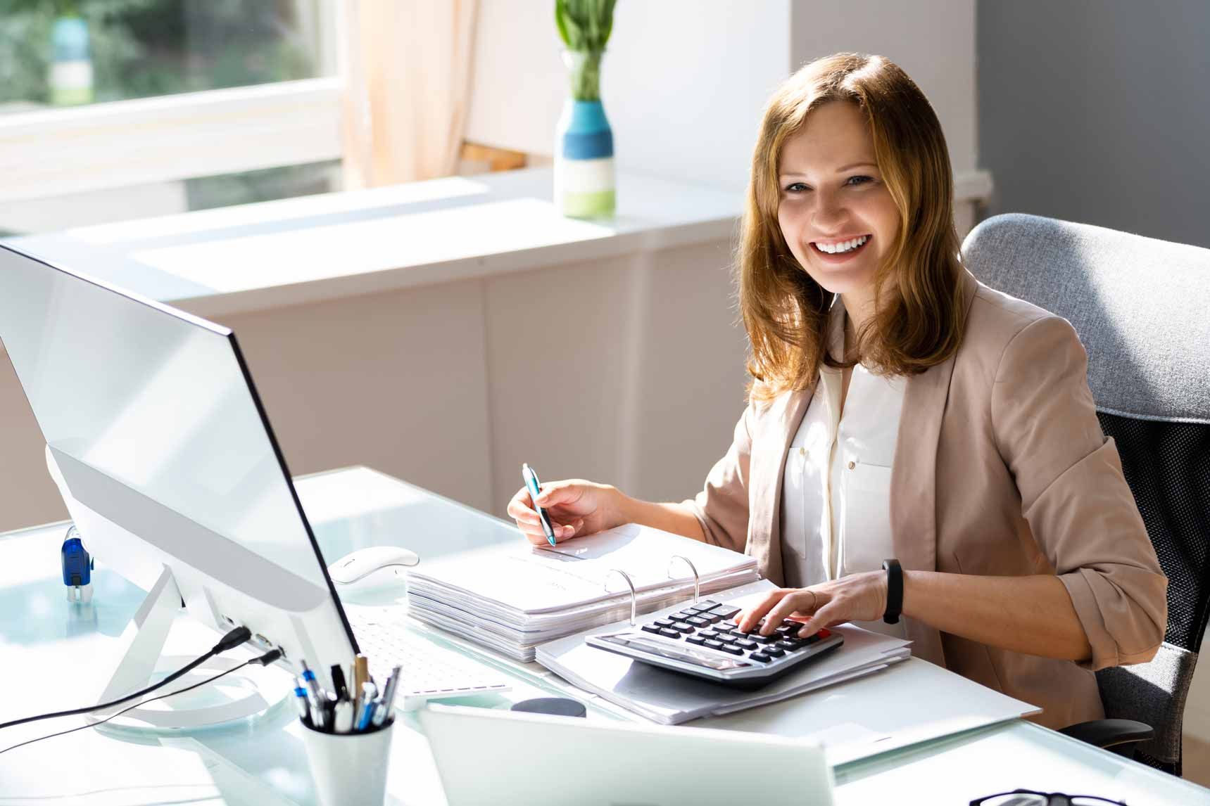 A smiling accountant working on tax paperwork with a calculator and a computer at her office desk.