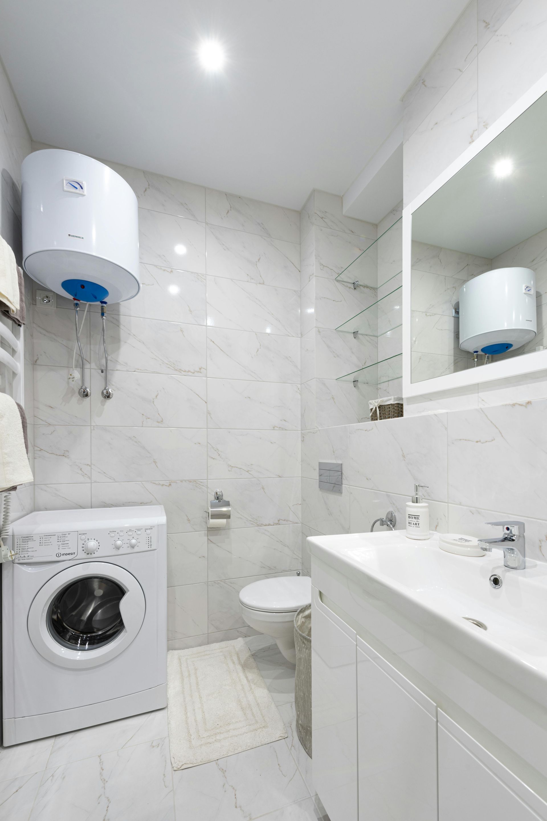 Small white bathroom with a washing machine, toilet, sink, mirrored cabinet, and water heater.
