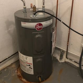Rusty Rheem water heater in a utility corner with pipes and a black hose attached