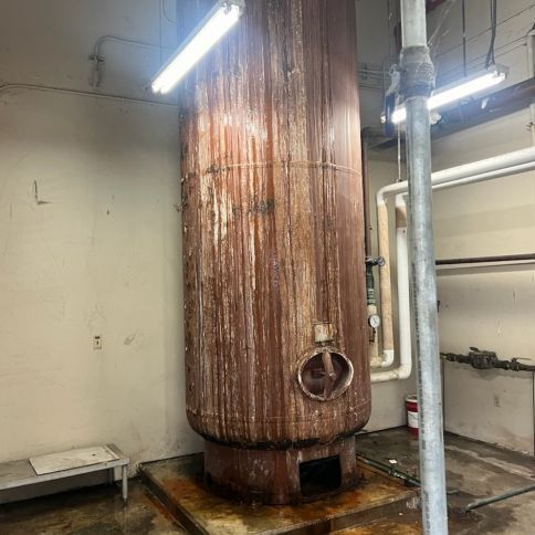 Rusty industrial tank in a utility room with pipes and fluorescent lights.