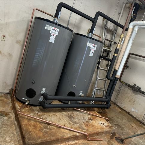 Two gray water heaters with black piping in a basement utility room
