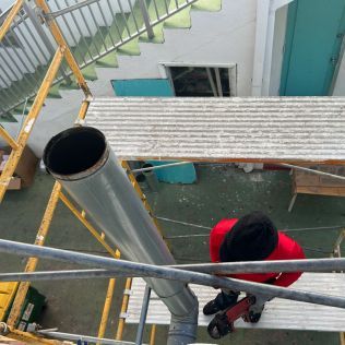 Worker in red jacket on a metal ladder beside a roof vent and corrugated roof panel