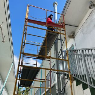 Worker on scaffolding beside a building, wearing a red jacket and hard hat.