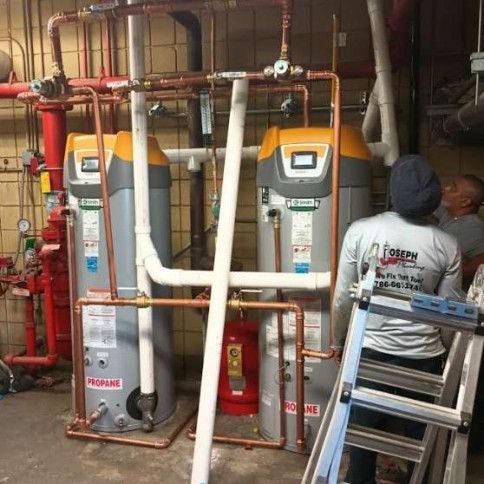 Two large HVAC units in a utility room, with white and red pipes and a worker on a ladder.
