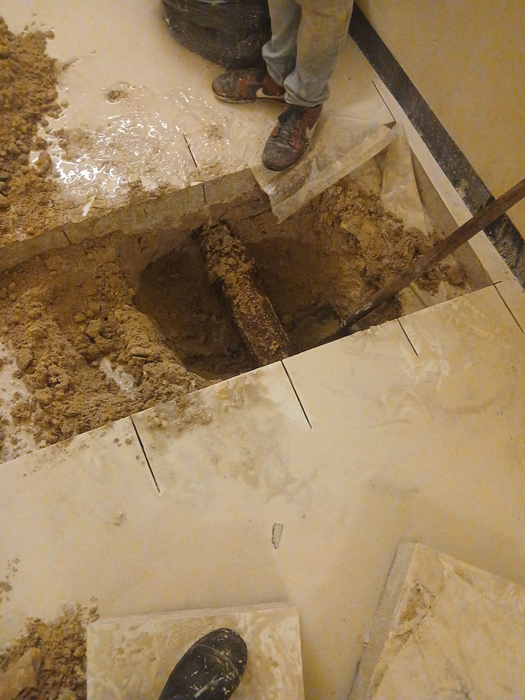 Two workers stand around a large square hole cut into a tiled floor, with dirt exposed beneath.