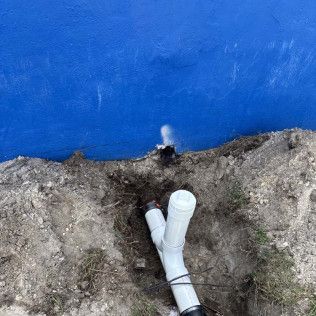 White PVC pipe emerging from dirt beside a blue wall.