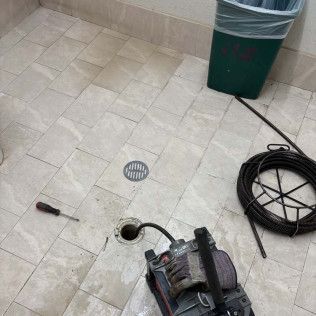 Small tiled floor with a drain, coiled black cable, green bin, and a portable machine near a hole.