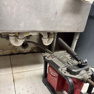 Red battery-powered floor scrubber under a stainless steel sink in a tiled utility room.