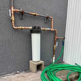 Copper pipes and filter tank beside a wall, with a green hose coiled on the ground.
