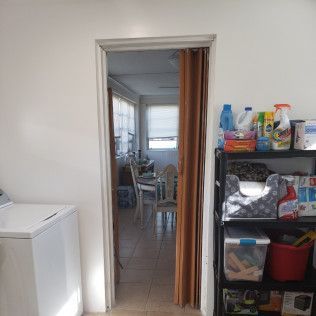 Laundry room with a washing machine, doorway to a dining area, and a shelf of cleaning supplies.