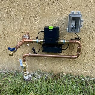Outdoor water filtration unit with copper pipes and a pressure gauge mounted on a stucco wall next to an outlet.