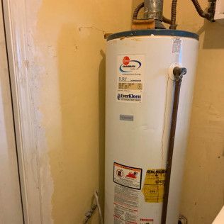White water heater in a closet corner with pipes and labels on a beige wall.