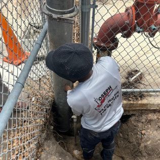 Worker in a hard hat inspecting a large pipe in a fenced industrial area with machinery nearby