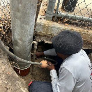 Worker in a gray hoodie repairing a pipe beside a chain-link fence and concrete wall.