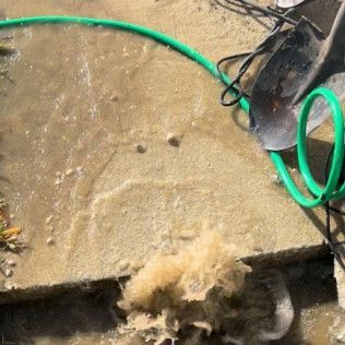 Floodwater gushing from a broken curb beside a green hose and black pump on wet concrete.
