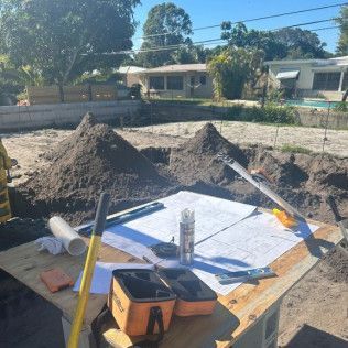 Surveying tools and plans on the ground beside dirt piles at a sunny construction site