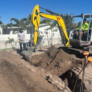 Yellow excavator digging a large trench on a sunny construction site, with workers nearby.