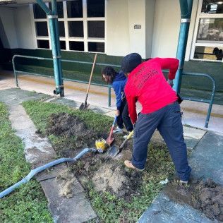 Two people repairing a broken water pipe near a building walkway