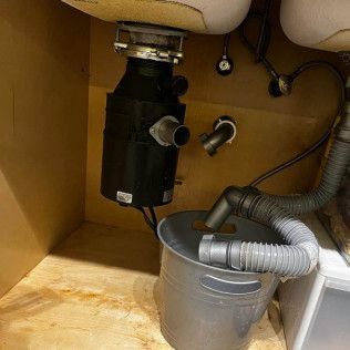 Under-sink plumbing with a black garbage disposal, gray hoses, and a small metal bucket collecting water.