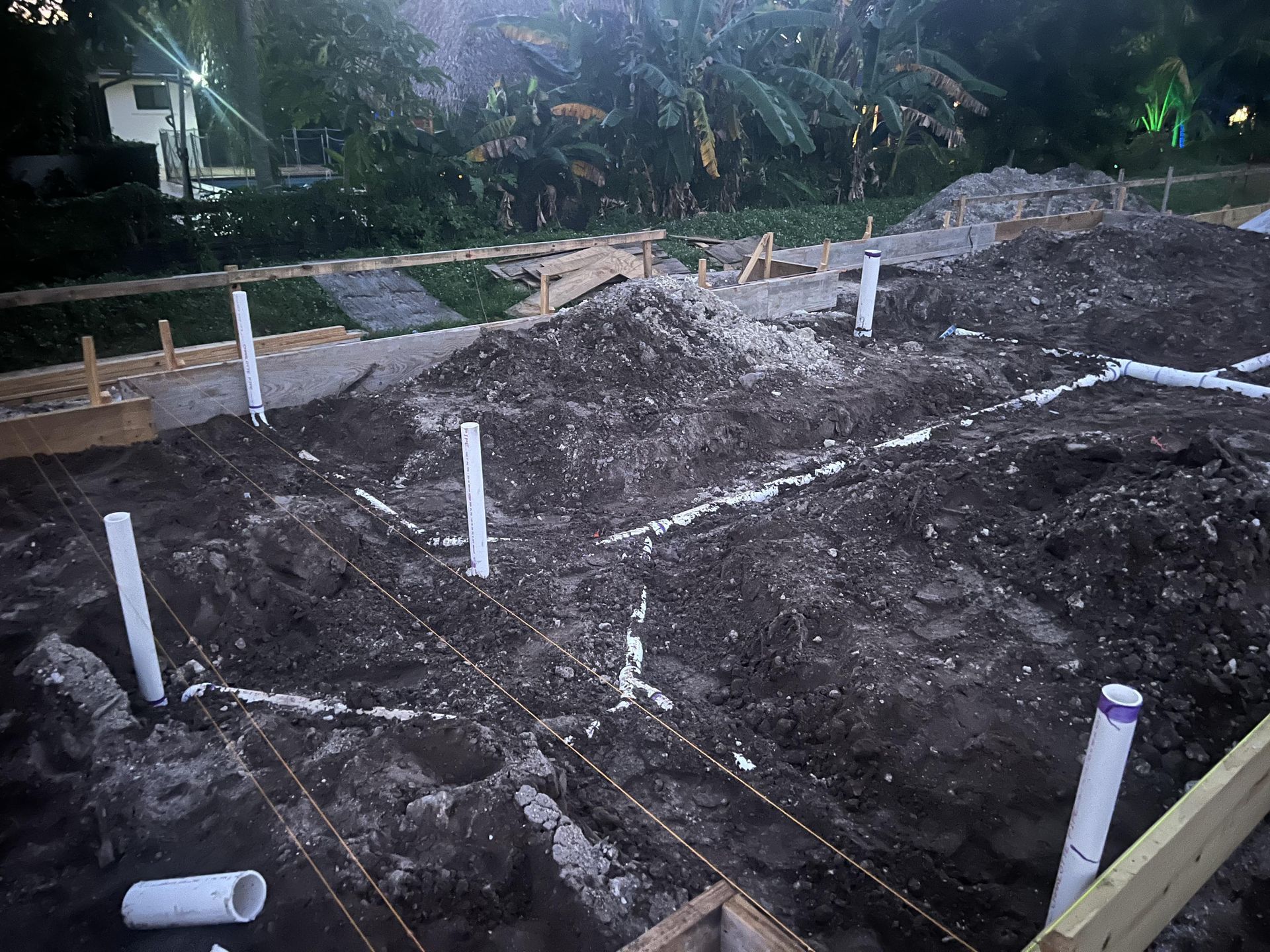 Cleared dirt lot with white survey stakes and string lines, next to a curb and tropical greenery.