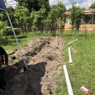 Trenched dirt path in a grassy yard with white PVC pipes and trees in the background