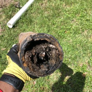 Gloved hand holding a rusted, corroded pipe end over grass, with a white pipe nearby.
