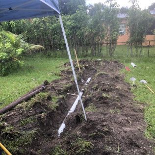 Trenched garden bed under a canopy tent beside a grassy yard, with white PVC pipes and piles of soil.