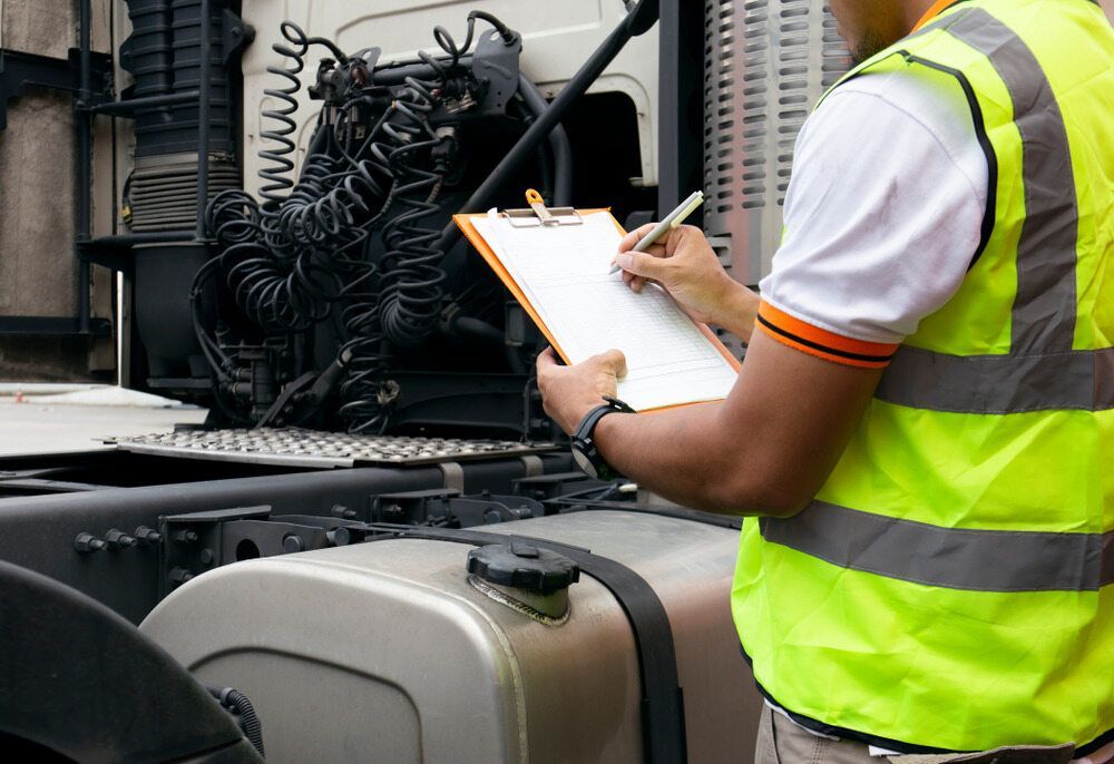 A Man in A Yellow Vest Is Writing on A Clipboard in Front of A Semi Truck — Coast Mechanical Services In Terrigal, NSW