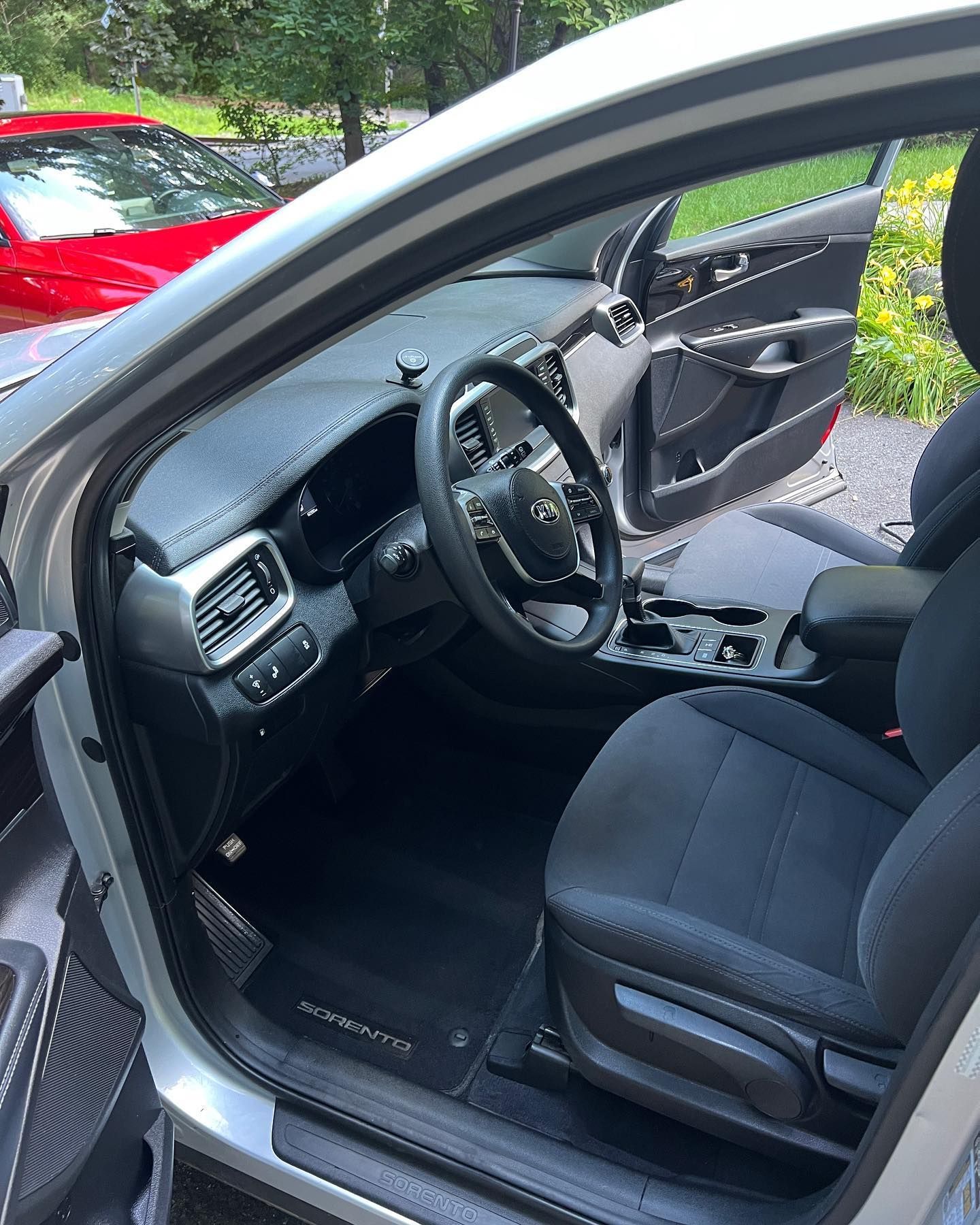 Car interior with driver’s door open, black seats and dashboard, parked outdoors near trees and a red car.