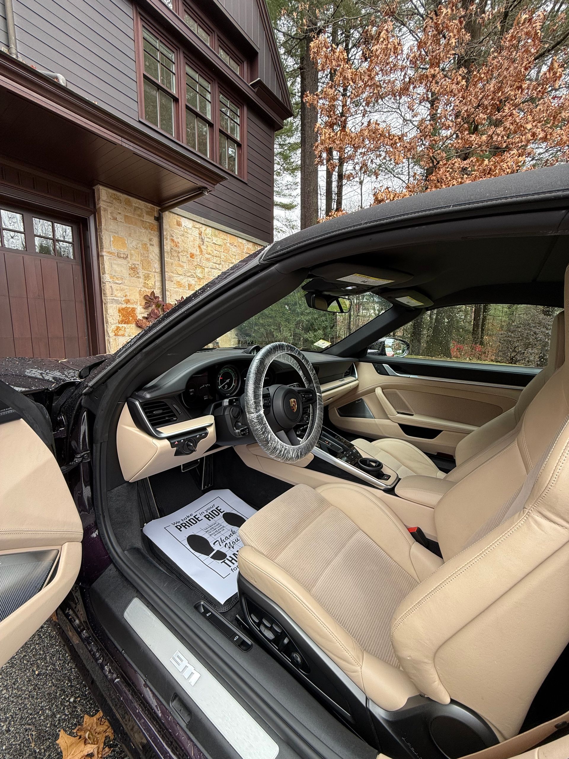 Open car door revealing beige leather interior and steering wheel beside a house in autumn.