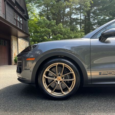 Gray Porsche SUV parked beside a house, showing gold rims and front side view.