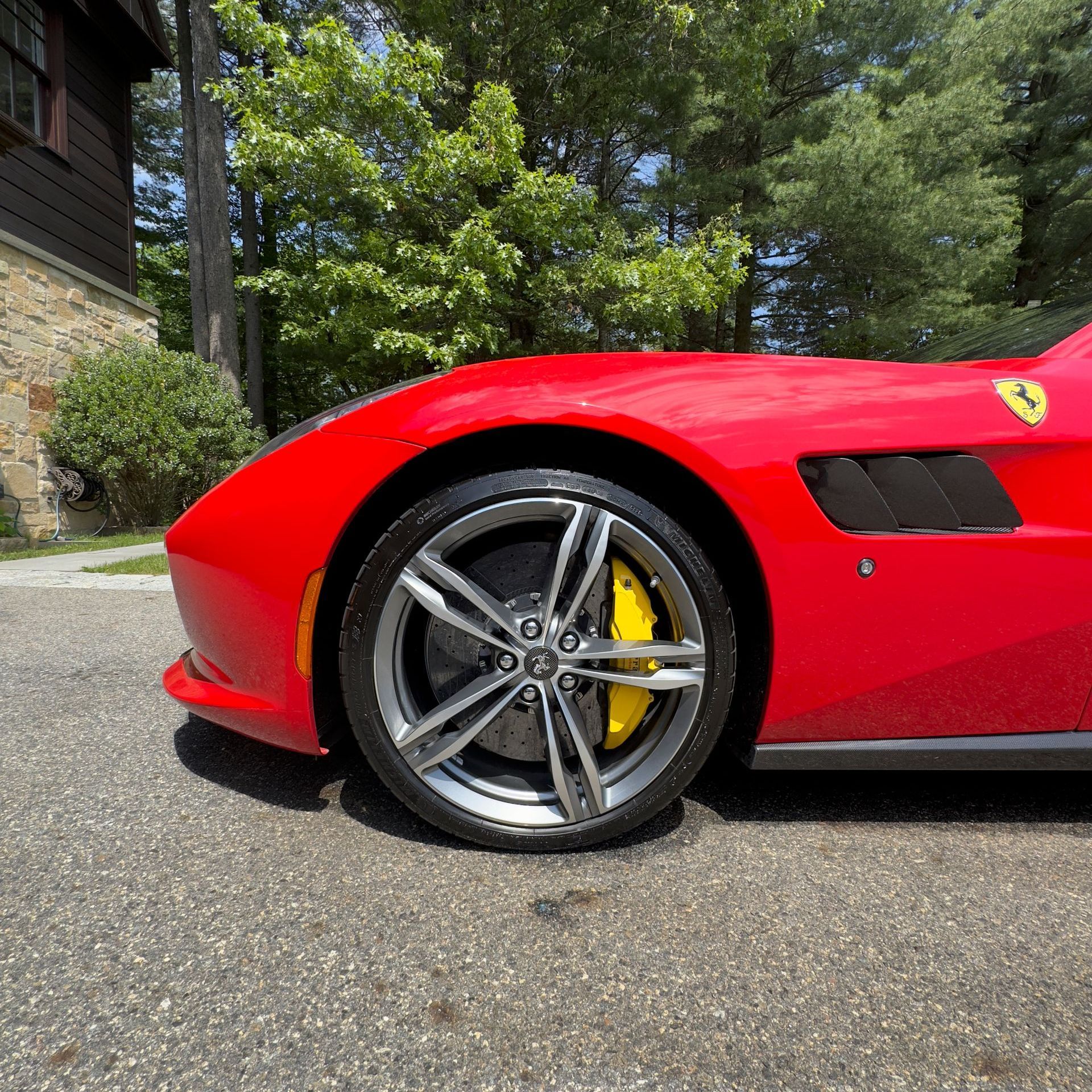 Red Ferrari sports car close-up, showing front wheel and yellow brake caliper on a sunny driveway.