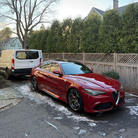 Red sports sedan parked on a snowy residential street beside a white van and hedge fence