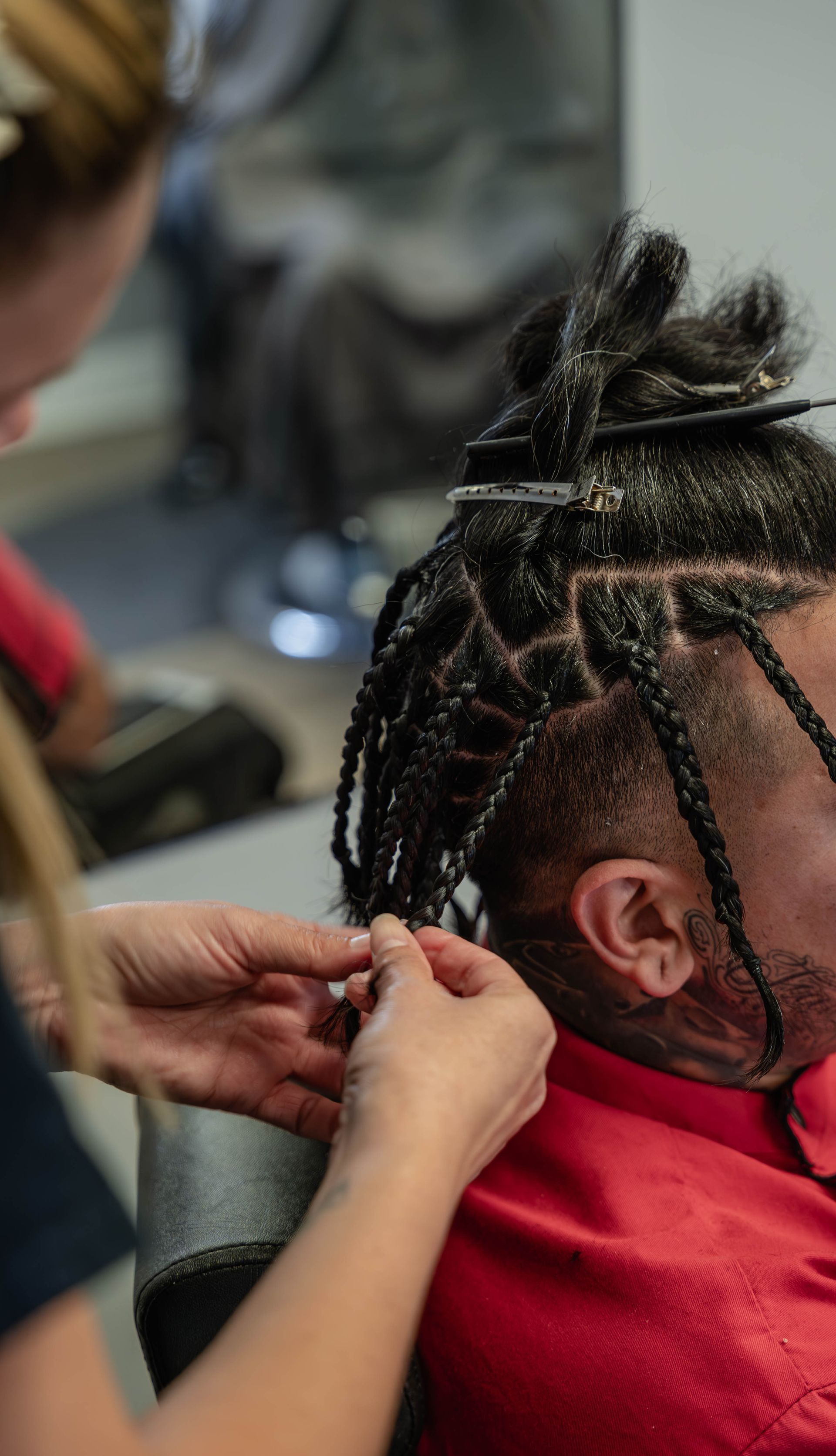 A stylist braids hair in small, box-section segments on a person wearing a red shirt. The Studio Barbershop
