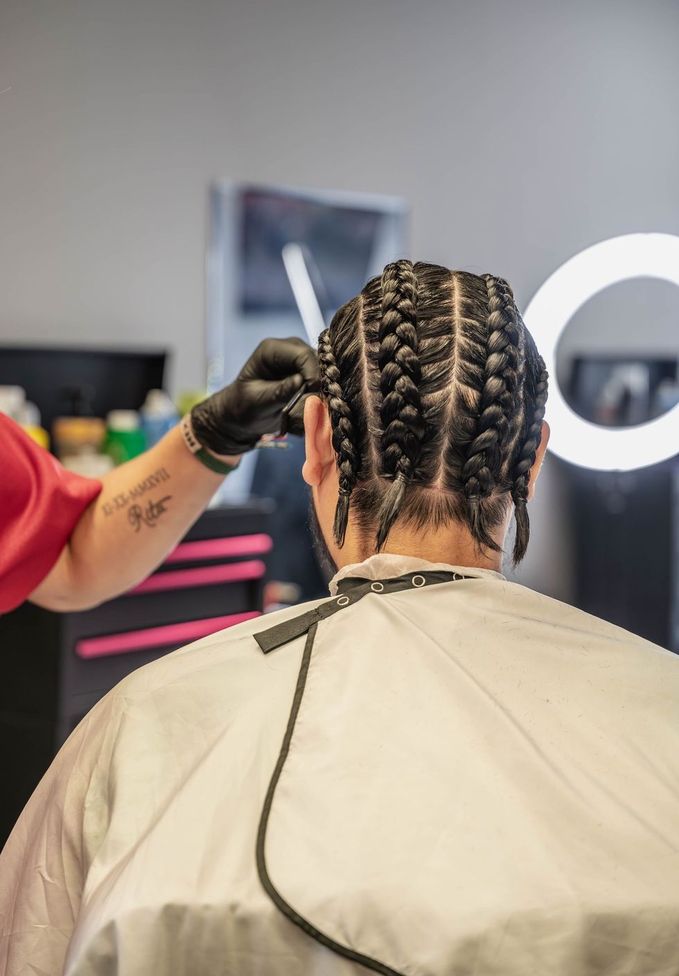 A barber wearing black gloves trims the back of a client's hair, which is styled in several neat cornrows. The Studio Barbershop