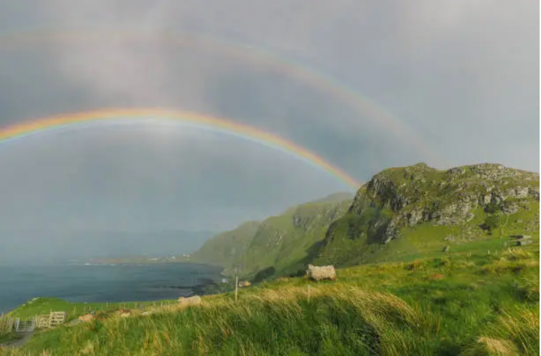 Double rainbow arches over a green, grassy cliffside overlooking the ocean; a cloudy sky.
