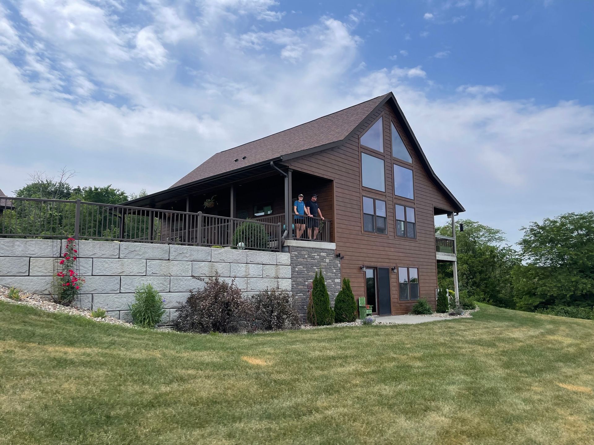 Brown building with a covered porch, large windows, and a retaining wall. Two people stand on the porch.