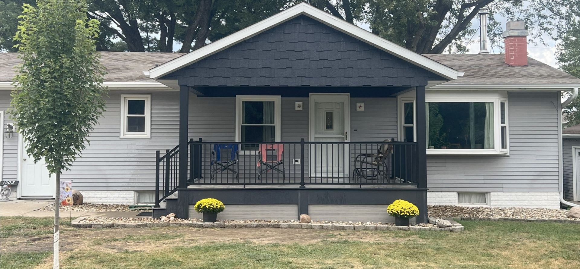 A gray ranch-style house with a black deck, small tree, and yellow flowers in front. Built in Center Point, Iowa