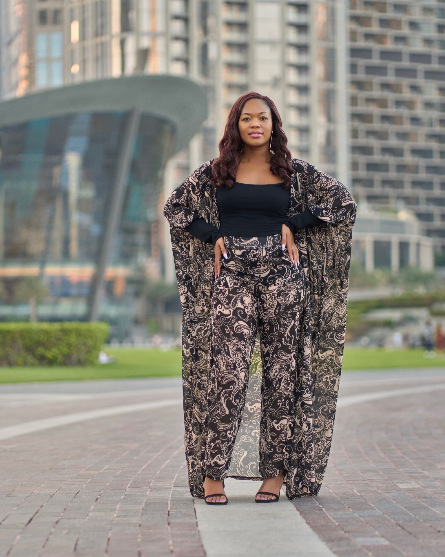Woman in black floral outfit standing on a paved walkway with city buildings in the background