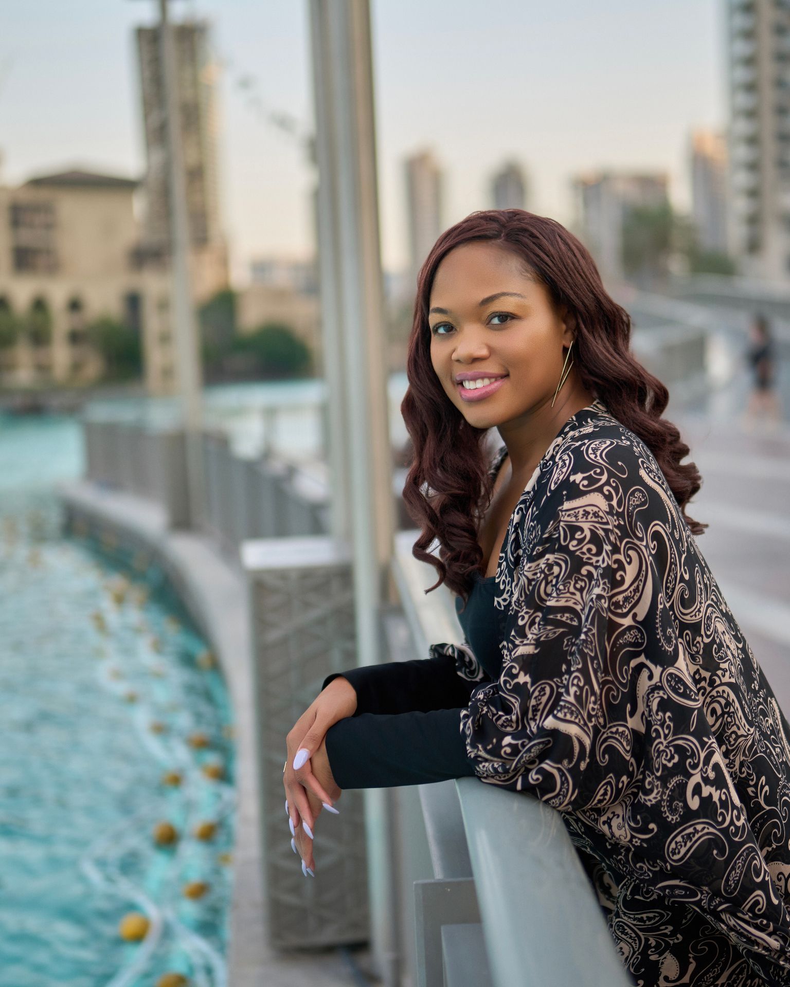 Smiling person in a patterned jacket leaning on a railing by a waterfront promenade at sunset
