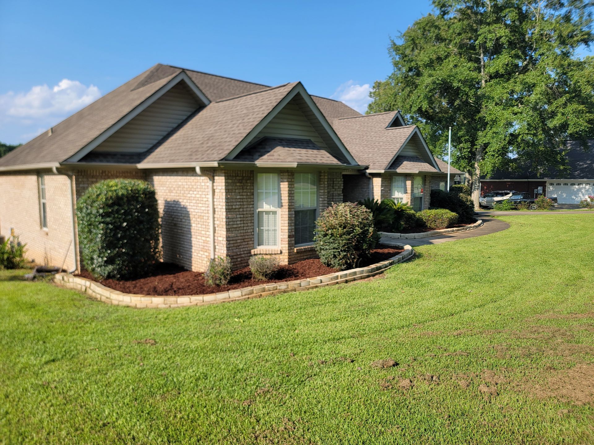 Single-story suburban house with brick exterior, green lawn, and trees under a blue sky